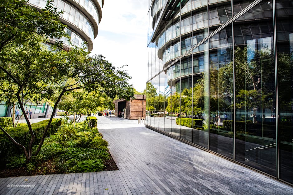A high-angle view of a modern urban rooftop garden on a multi-storey building. The garden features several narrow, elongated planting beds filled with soil and small plants, bordered by glass railings